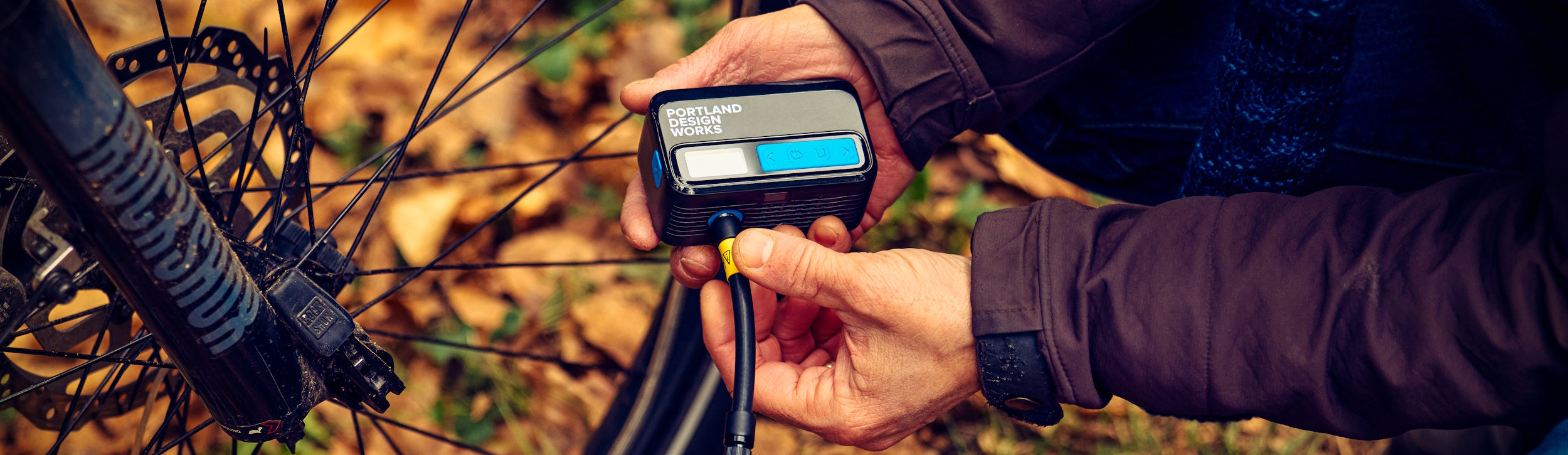 A rider inflating a mountain bike tire with an Essentials Electric Bike Pump. Leaves can be seen on the ground.
