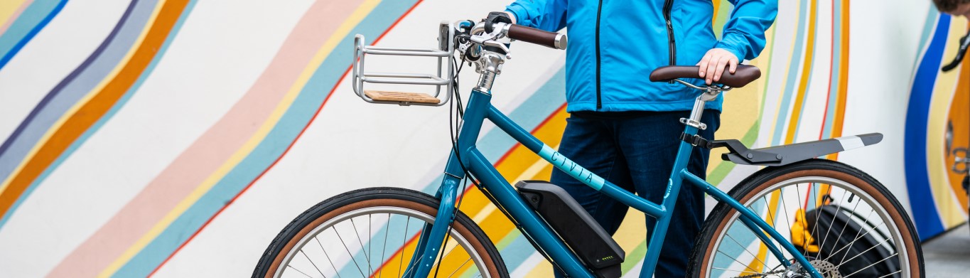 A blue e-bike with battery mounted on the downtube with a PDW Task Basket on the handlebars and PDW Origami fender mounted to the seatpost. A rider in a light blue rain jacket is holding the bike.