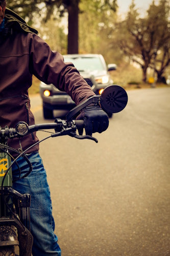 Watch Your Back XL bicycle mirror on a handlebar of a bike with the back of the mirror visible and a truck coming up behind the bicycle rider.